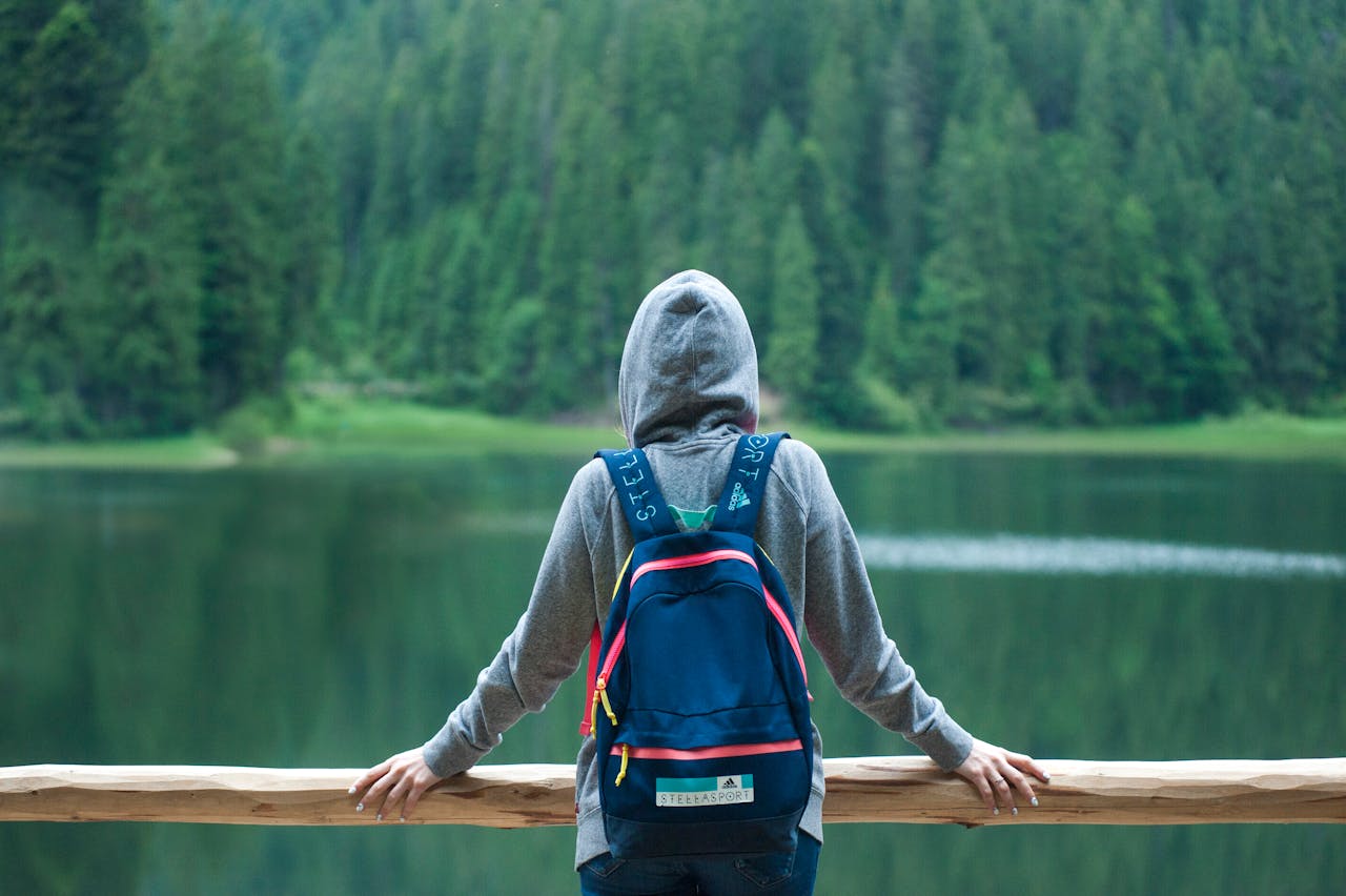 gallery-01 A person in a hoodie stands by a serene lake surrounded by lush greenery, enjoying the peaceful outdoors.